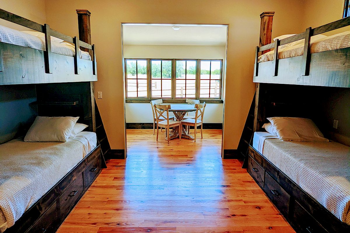 The image shows a room with two double bunk beds, hardwood floors, a ceiling fan, and a small table in a windowed area at the far end.
