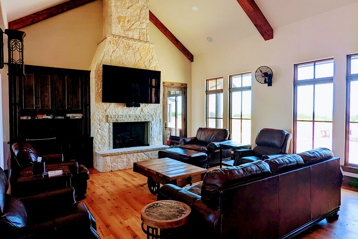 A spacious living room with leather sofas, a stone fireplace, wooden floors, high ceiling with beams, and large windows for natural light.