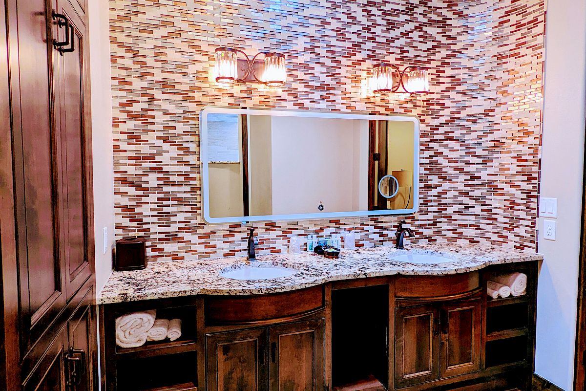 A bathroom with wooden cabinets, a patterned tile wall, a large mirror, double sinks, and two wall lights.