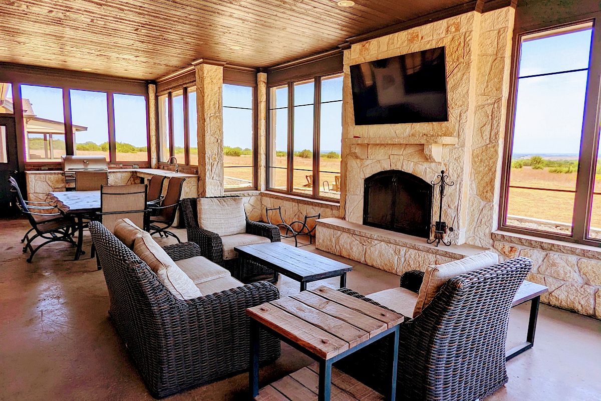 A cozy indoor lounge with wicker furniture, a fireplace, and a TV. Large windows bring in natural light, creating a relaxed atmosphere.