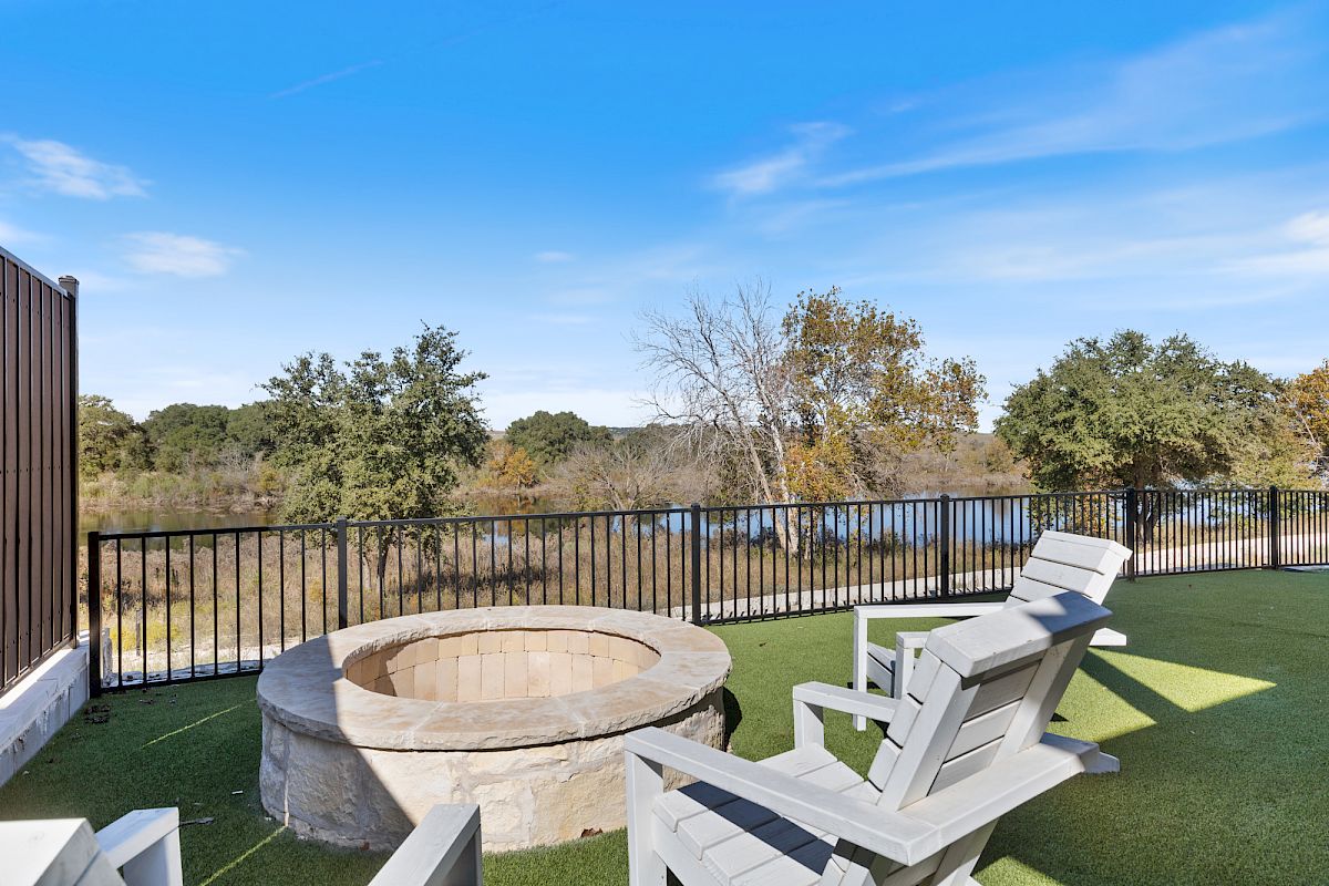 The image shows a patio with Adirondack chairs, a fire pit, and a view of trees and a body of water behind a black metal fence.
