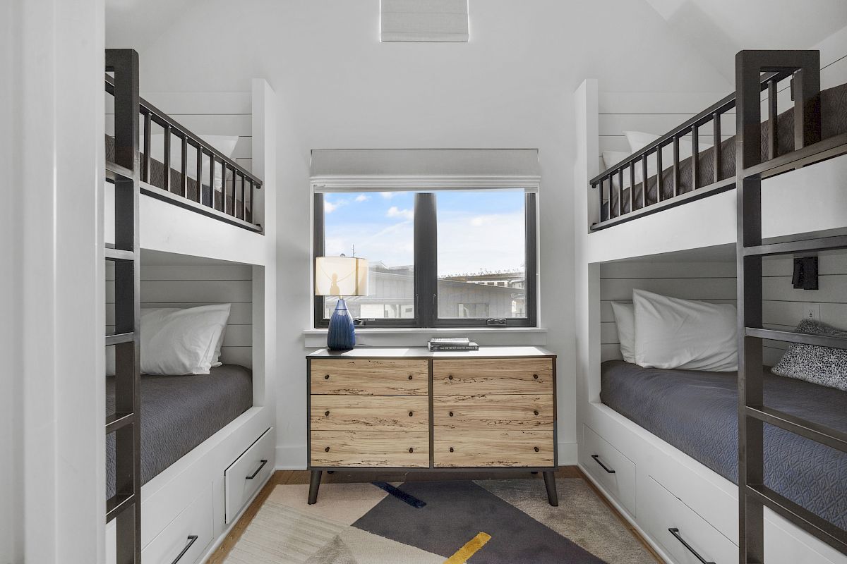 The image shows a room with two bunk beds and a wooden dresser between them, under a window with blinds. A lamp is on the dresser.