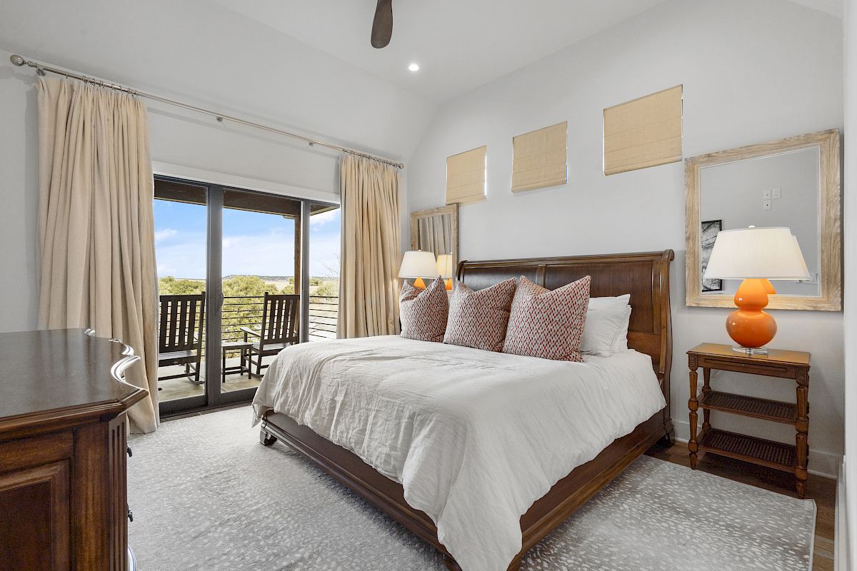 A cozy bedroom with a large bed, orange lamps, and a view of a balcony. Neutral tones and ample natural light enhance the calm vibe.