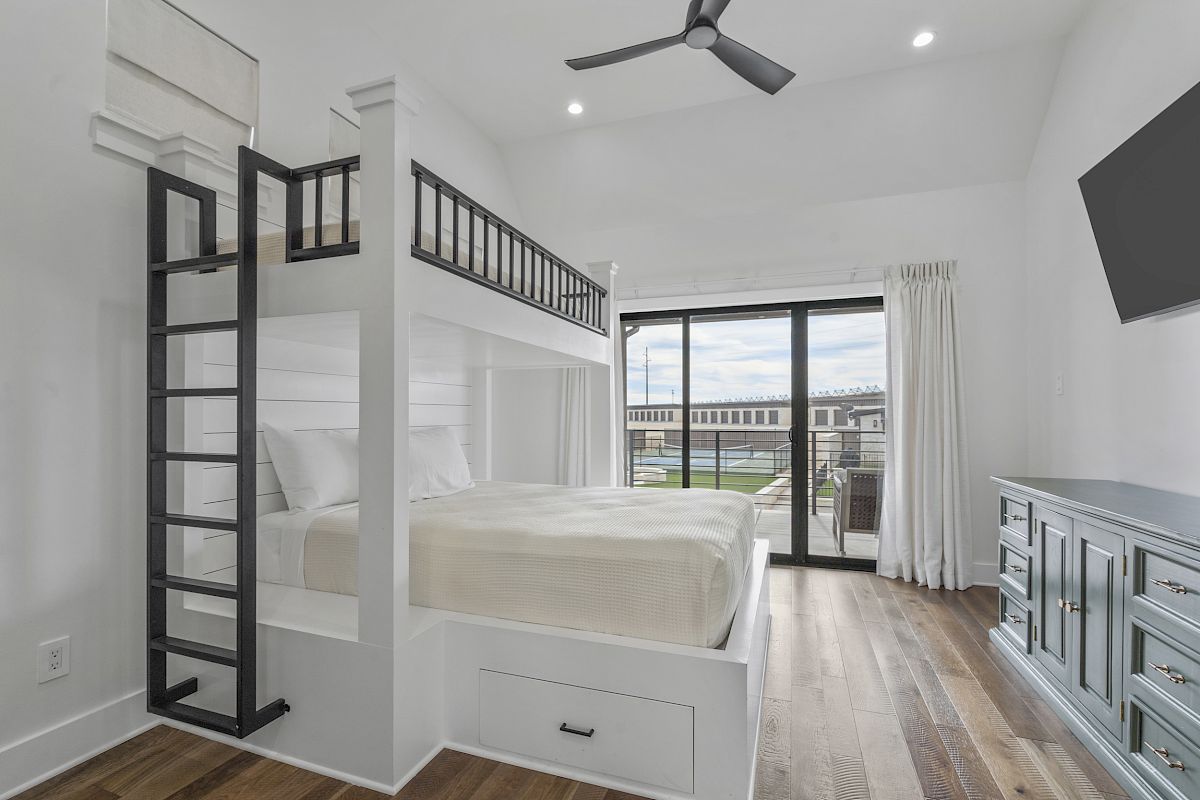 A modern bedroom with a bunk bed, wood flooring, a TV, and a dresser, featuring large sliding glass doors leading to a balcony.