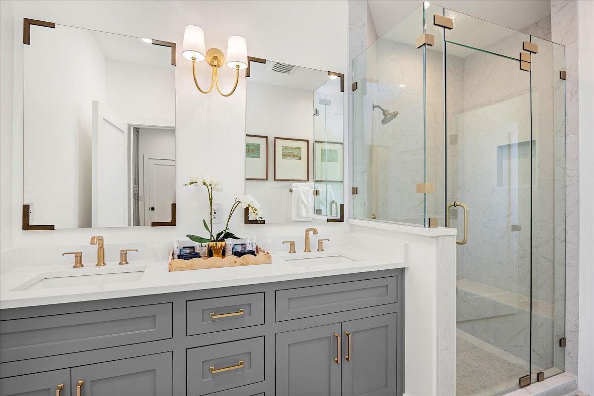 A modern bathroom with a gray double vanity, gold fixtures, large mirrors, glass shower, and wall sconces for lighting.
