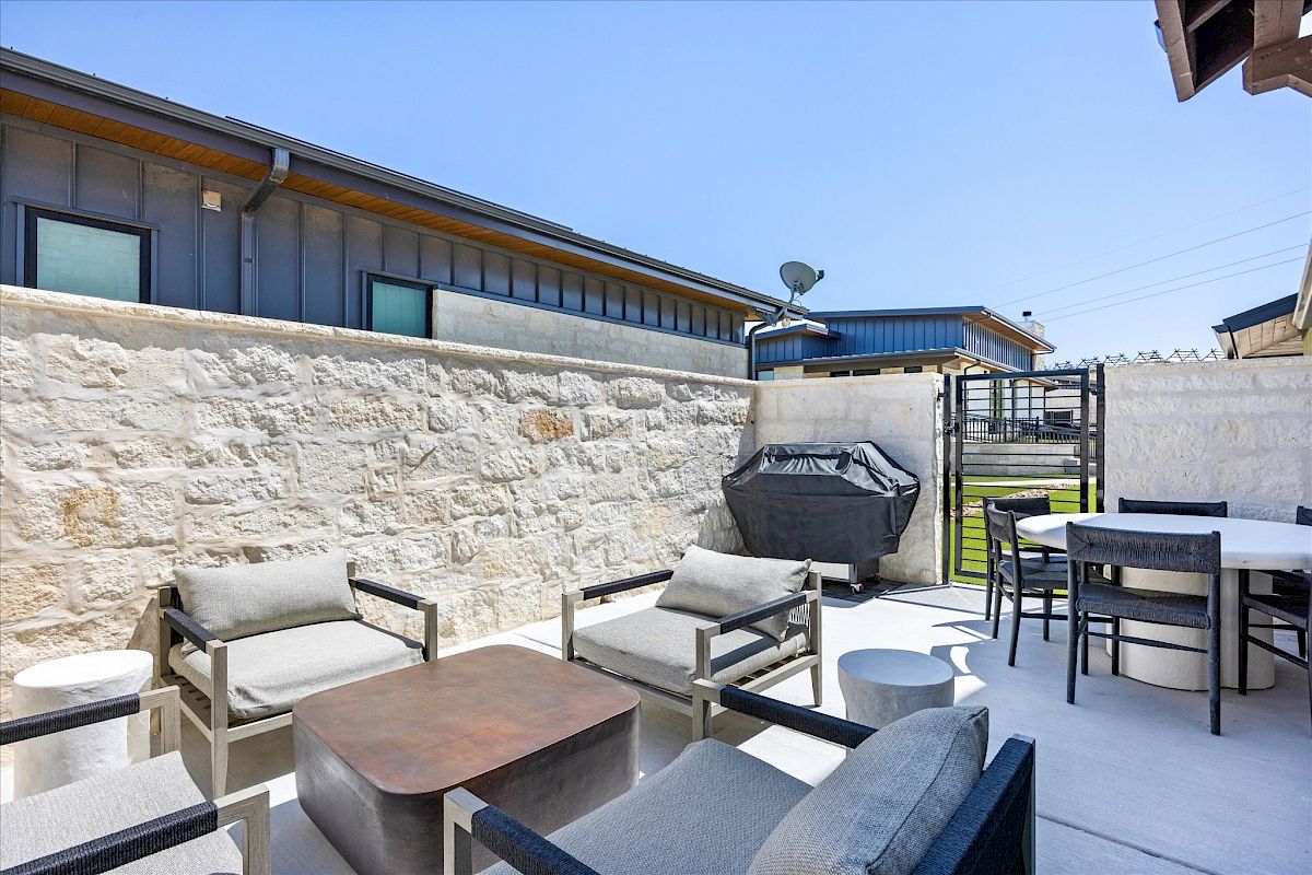 A modern outdoor patio with cushioned seating, a wooden table, a grill, and a dining set, enclosed by stone walls.
