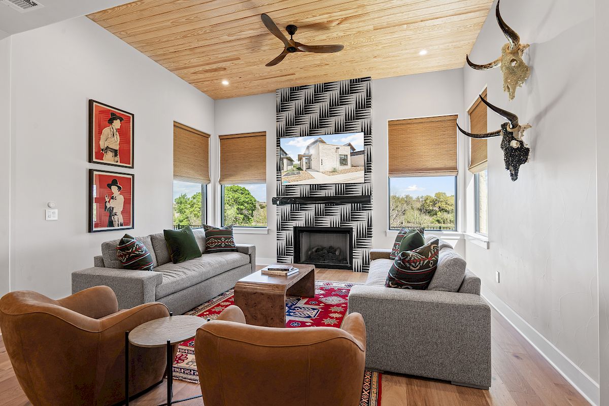 The image shows a modern living room with gray sofas, leather chairs, decorative art, a patterned fireplace, and large windows with blinds.
