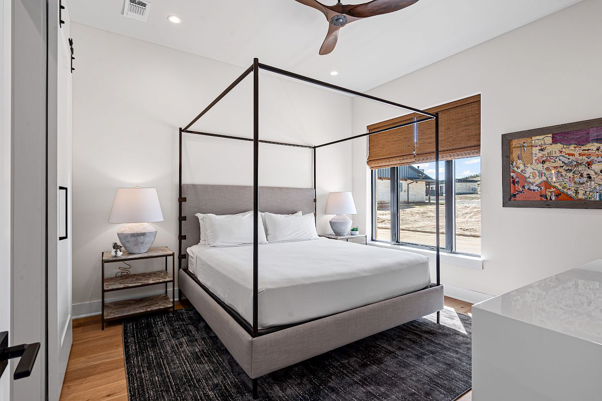 The image shows a modern bedroom with a canopy bed, lamps, artwork, and a window with blinds, creating a stylish and cozy ambiance.