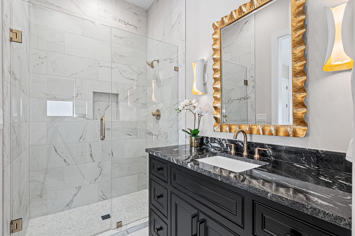 A modern bathroom with a glass shower, marble tiles, black vanity, gold-framed mirror, and elegant lighting fixtures, ending the sentence.