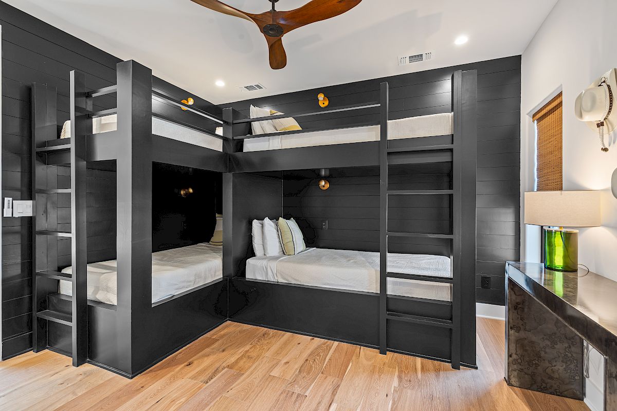 The image shows a room with modern black bunk beds, wooden floor, ceiling fan, and a lamp on a dresser.