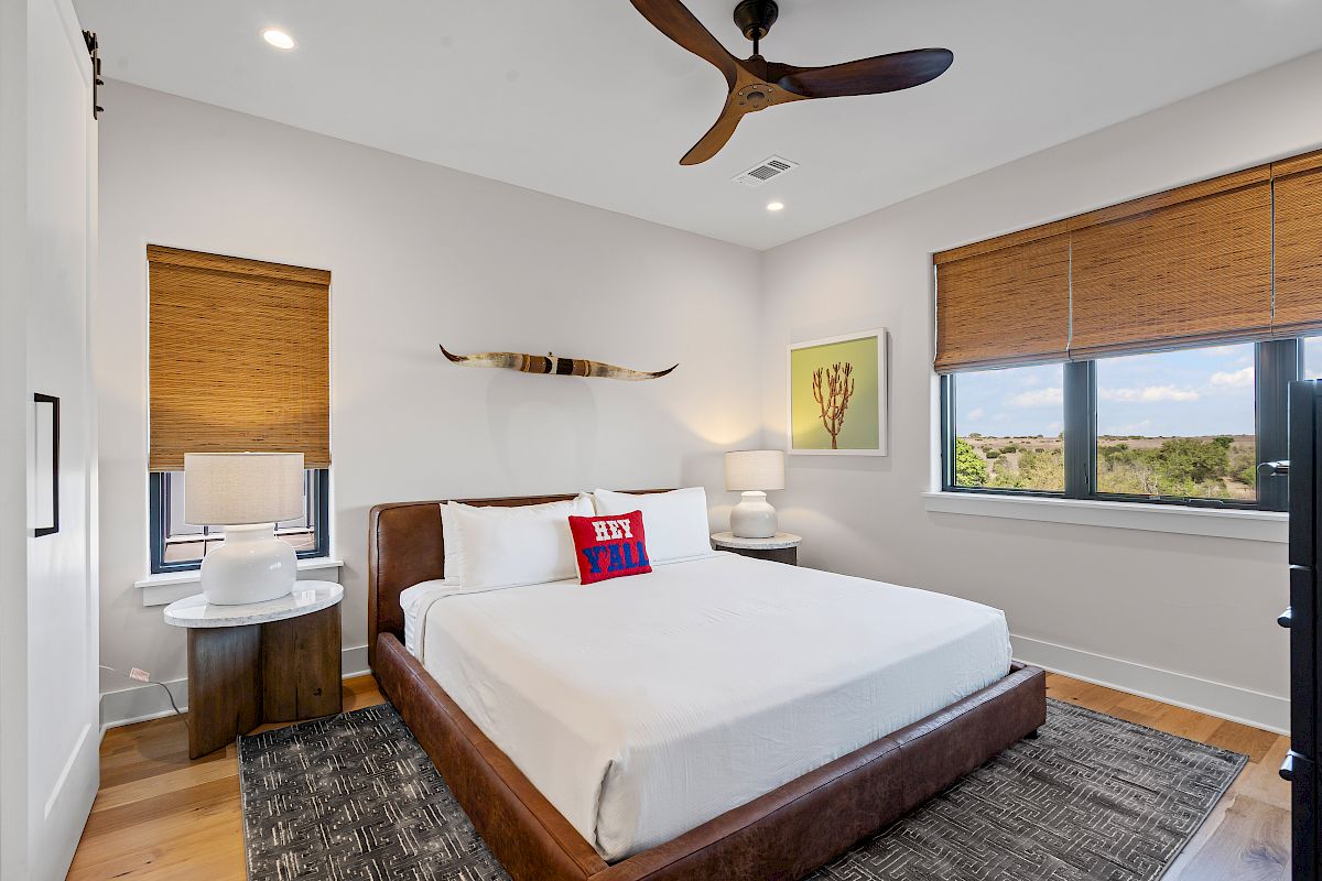 A cozy bedroom with a double bed, wall decor, two side tables, lamps, and large windows with blinds.