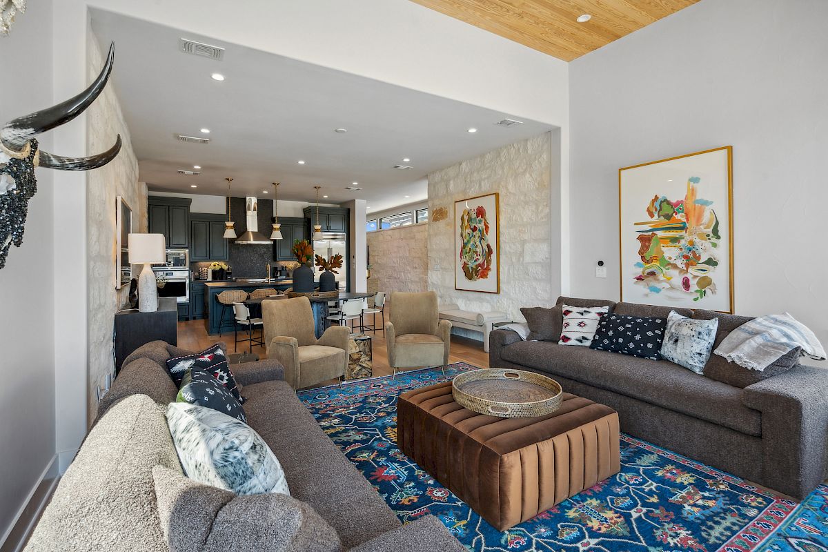 A modern living room with gray sofas, a blue patterned rug, art on walls, and a view into the kitchen area with pendant lights.