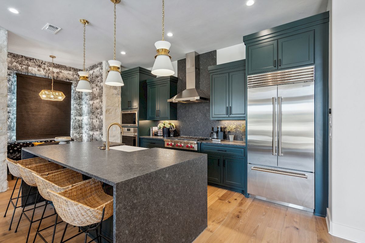 A modern kitchen with a large island, wicker stools, pendant lights, dark green cabinets, and stainless steel appliances.