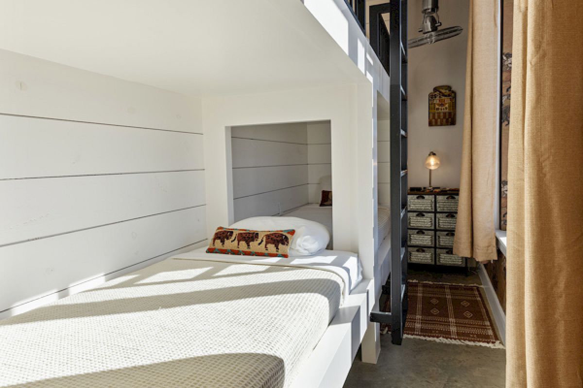 A cozy bunk bed setup with a rug, pillow, and decorative elements in a narrow, well-lit room with light curtains and industrial touches.