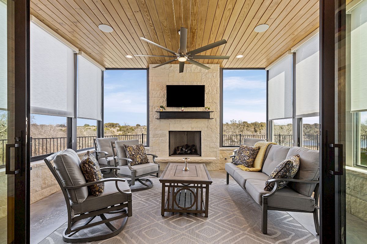 A cozy enclosed patio featuring a ceiling fan, a stone fireplace with a TV, cushioned seating, and large windows with blinds.