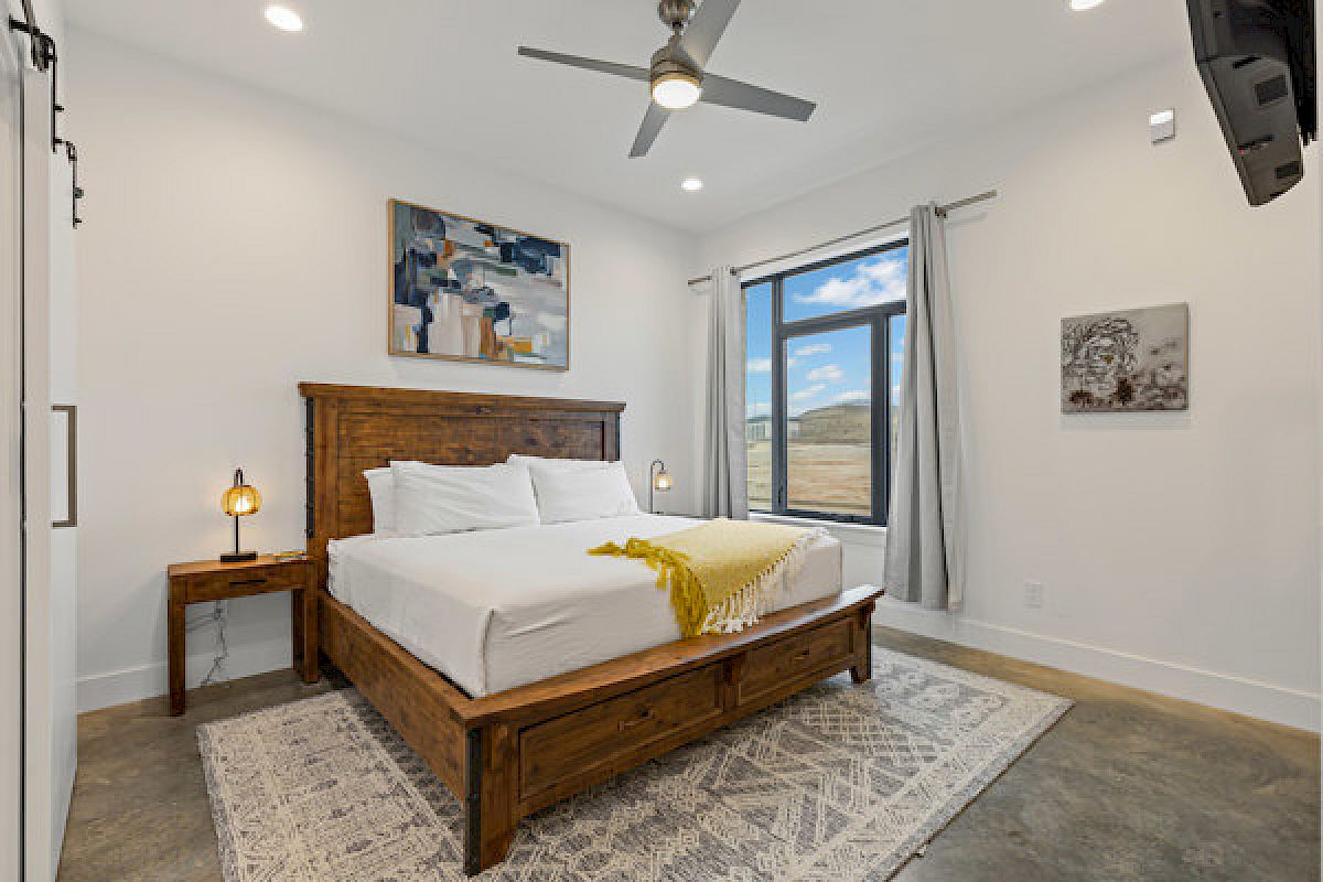 A bedroom with a wooden bed, white bedding, yellow throw, art on walls, large window, ceiling fan, and a wall-mounted TV.