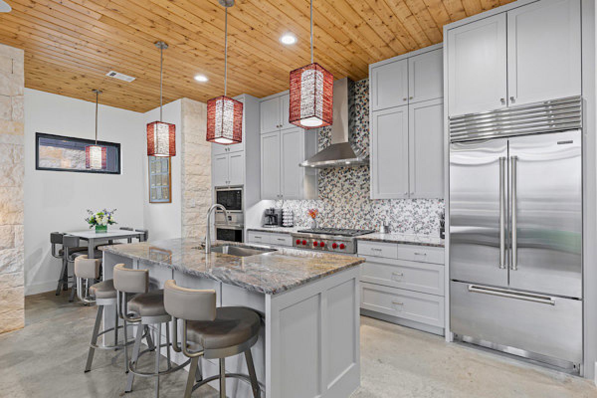 A modern kitchen with a marble island, gray cabinets, red pendant lights, and a stainless steel refrigerator and appliances.