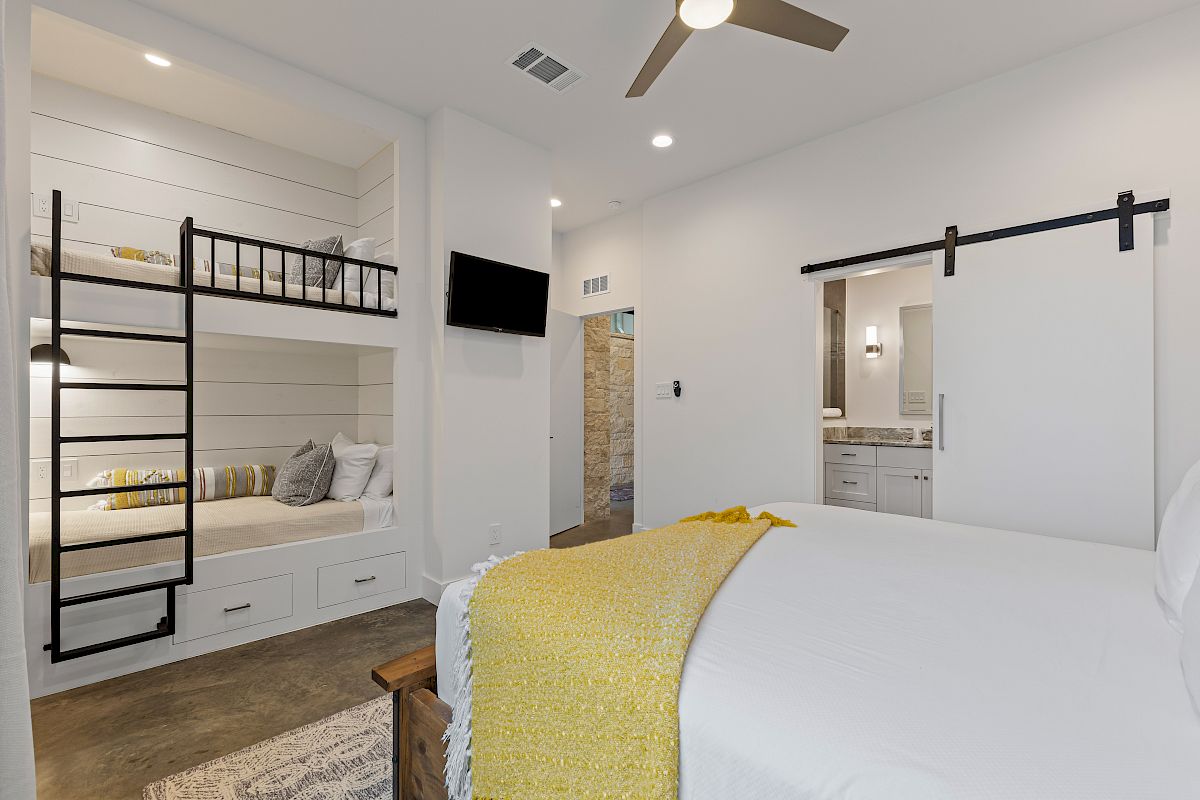 The image shows a modern bedroom with a bunk bed, a bed with a yellow throw, a ceiling fan, a TV, and a sliding door leading to a bathroom.