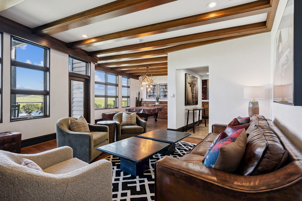 A modern living room with a leather sofa, armchairs, large windows, a patterned rug, and wooden ceiling beams, overlooking countryside views.