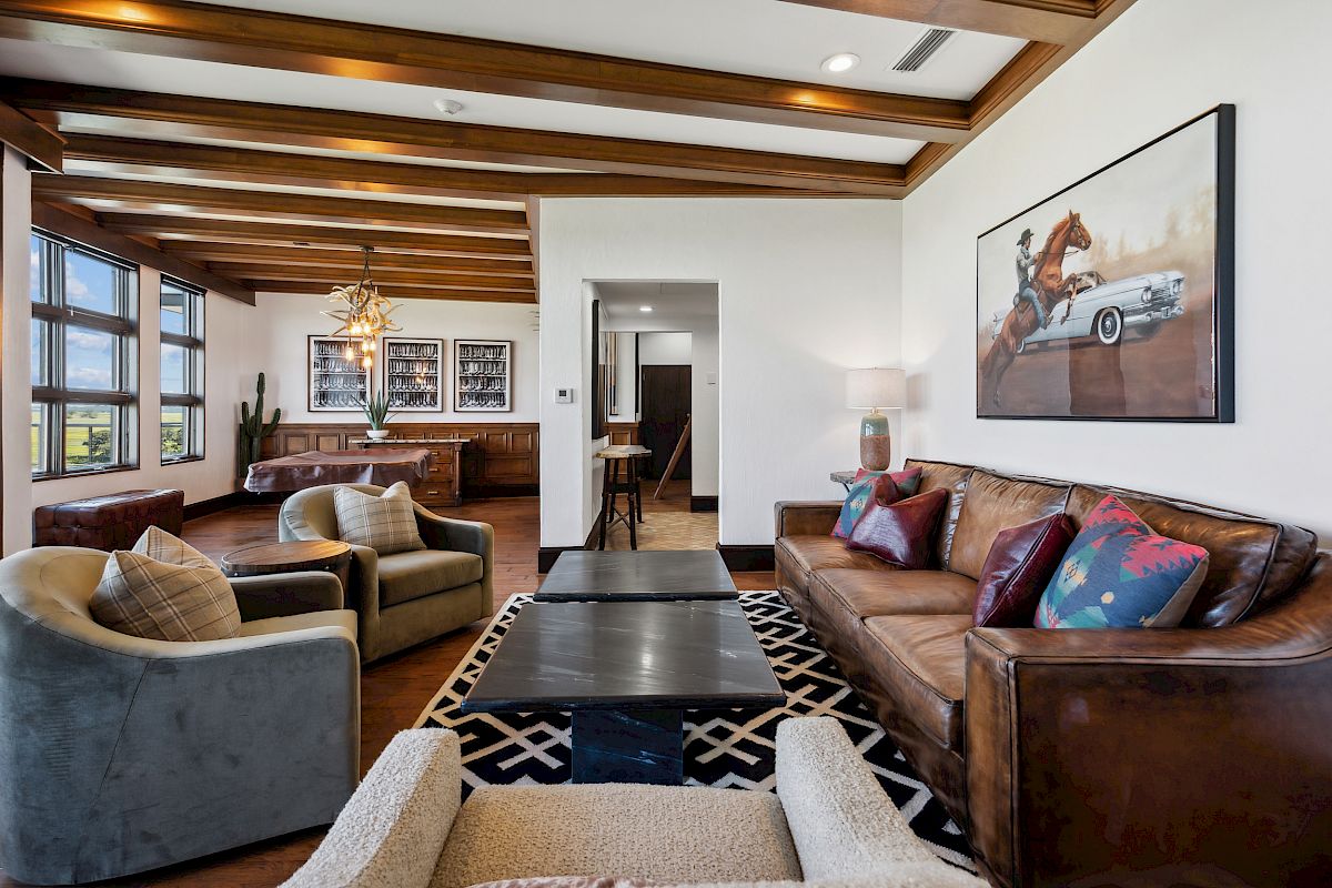 A modern living room with leather sofas, patterned rug, and horse artwork. Wooden beams and a dining area are visible in the background.
