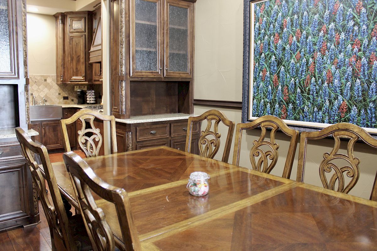 Dining room with a wooden table, chairs, a kitchen in the background, and a vivid floral painting on the wall.