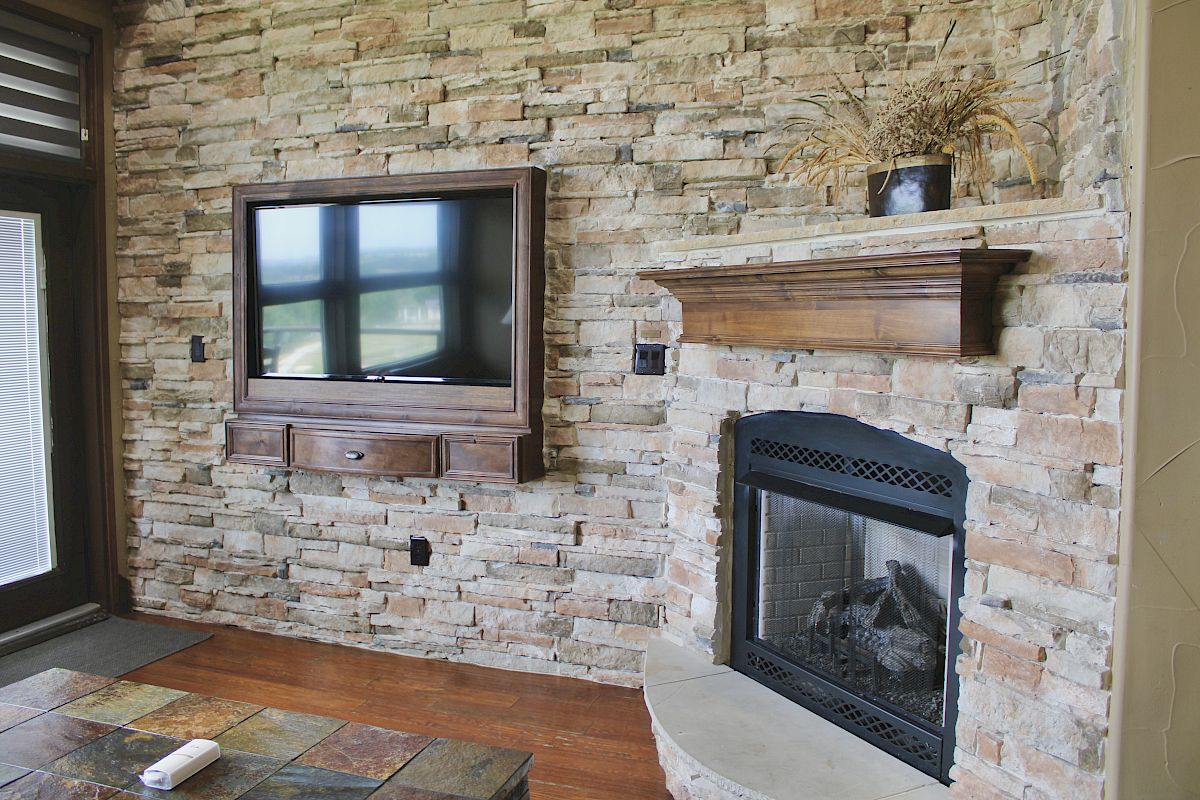A room with a stone wall features a mounted TV, a fireplace, and a wooden mantle with a plant on top.