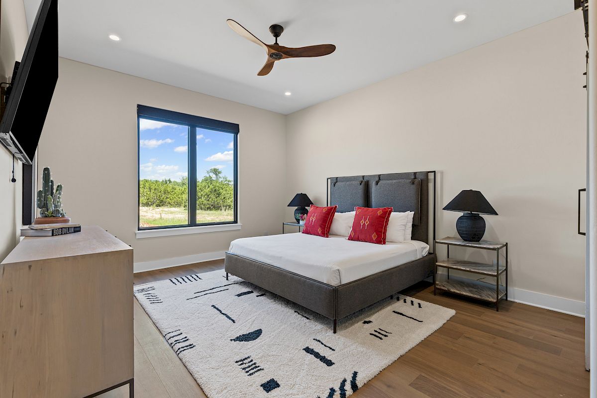 A modern bedroom with a large bed, red pillows, a wall-mounted TV, a ceiling fan, and a window showing a scenic view.