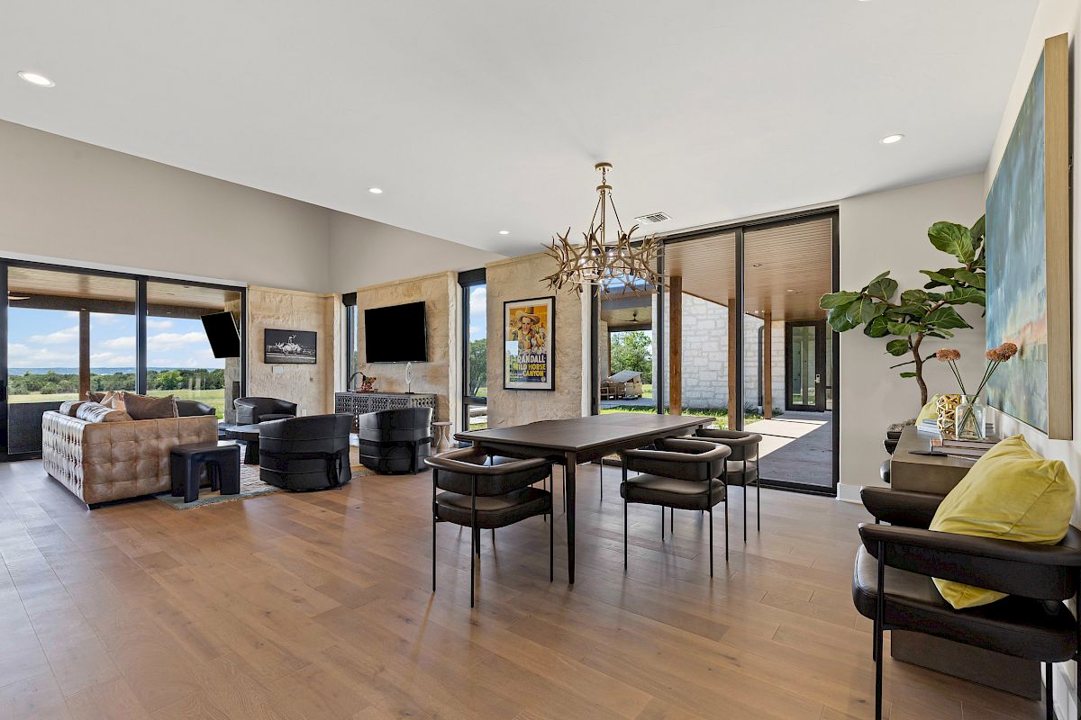 A spacious living area with modern furniture, large windows, a dining table, and a chandelier, offering a view of the outdoors.