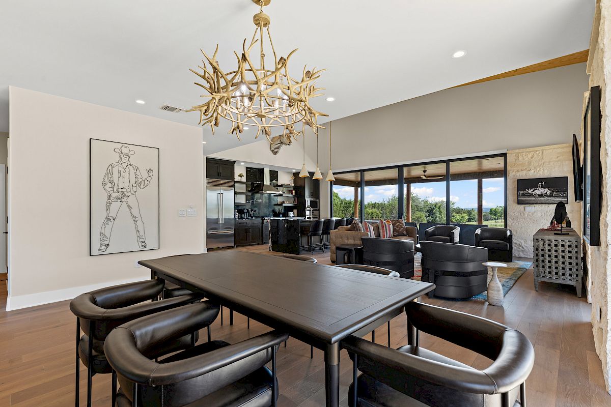 A modern living area with a dining table, antler chandelier, large windows, black chairs, and cowboy art on the wall.