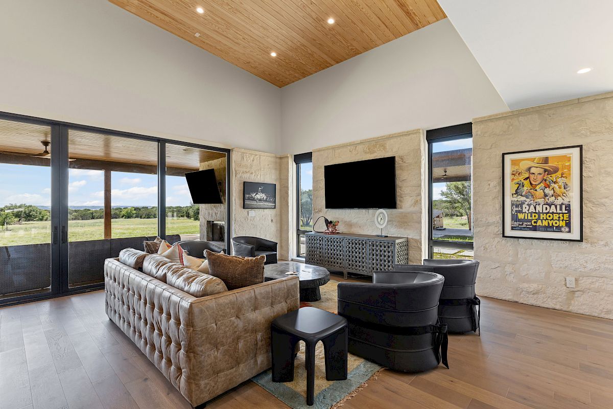 A modern living room with a leather sofa, armchairs, TV, and poster on the wall. Large windows offer a view of the outdoors.