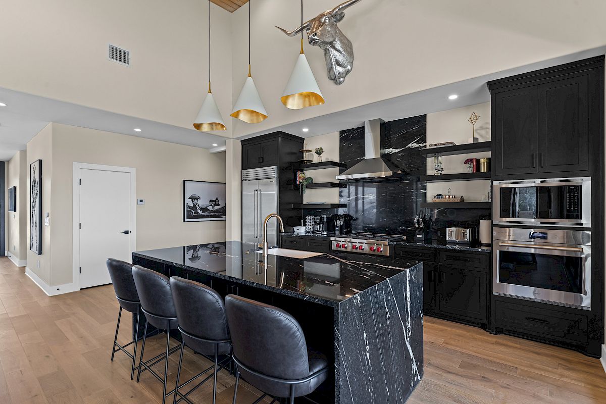 A modern kitchen with a black marble island, bar stools, pendant lights, stainless steel appliances, and a metallic deer head decoration.
