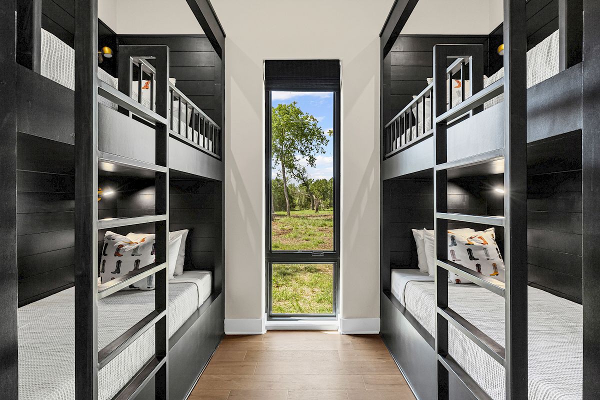 The image shows a room with four dark wooden bunk beds, pillows with patterns, and a window overlooking trees and grass.
