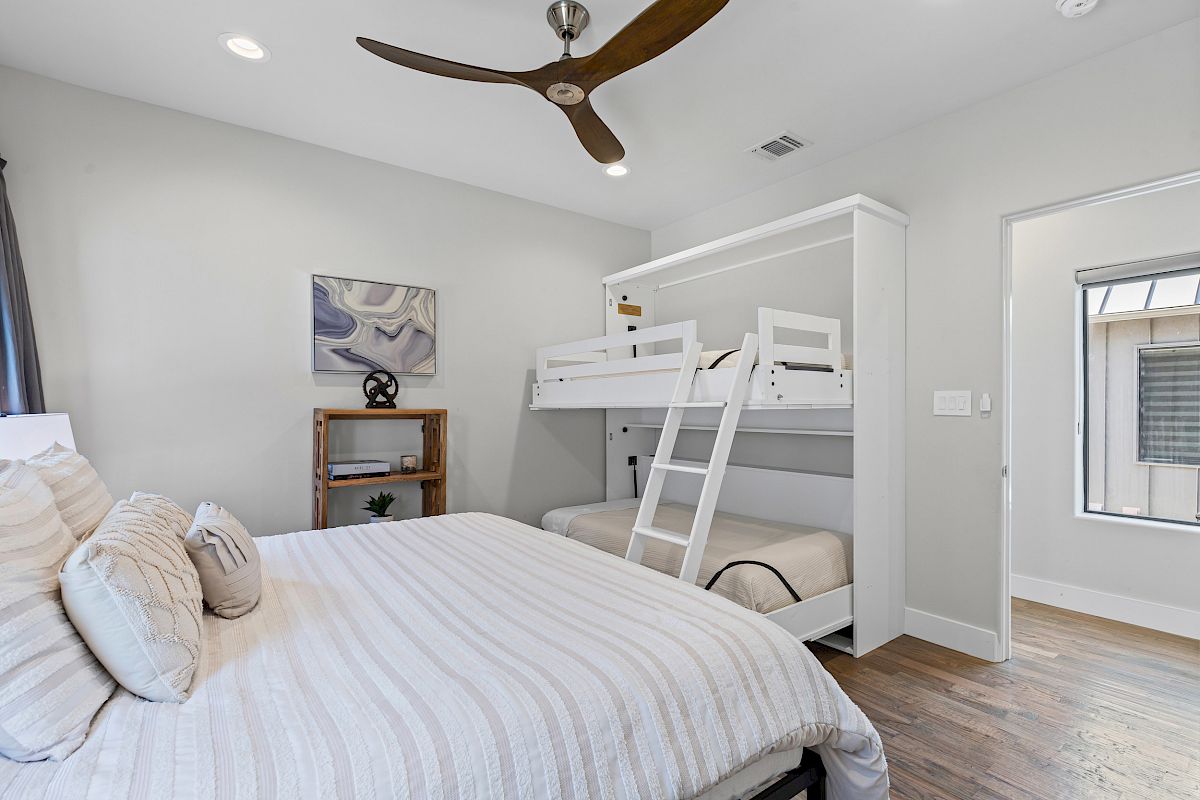 A bedroom with a double bed, bunk beds, wooden flooring, ceiling fan, bookshelf, and artwork on the wall.