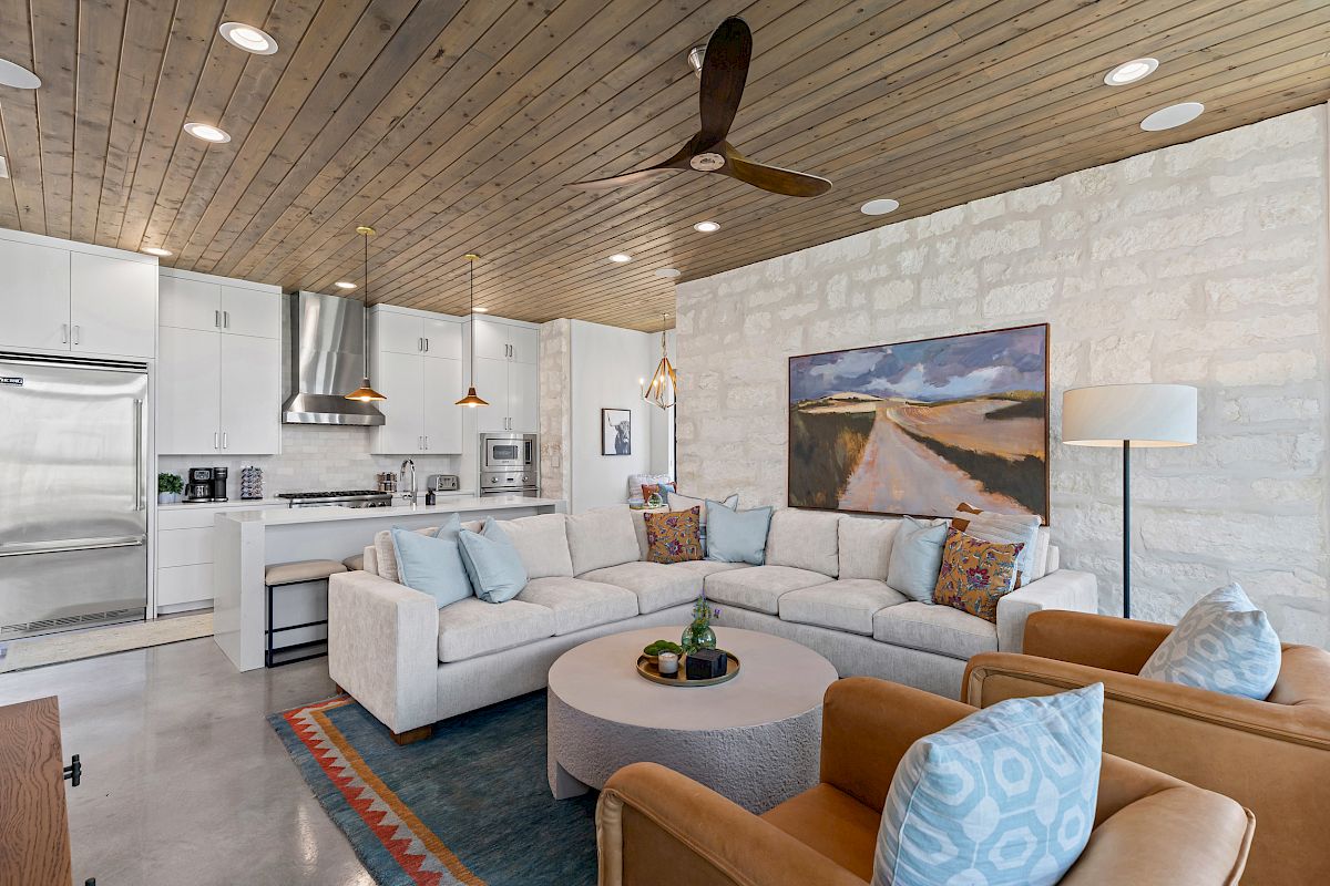 A modern living room with a large sectional sofa, kitchen area, wooden ceiling, ceiling fan, and artwork on a stone wall.
