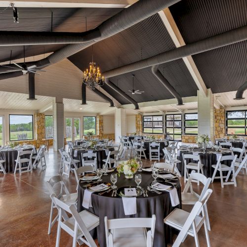 The image shows a decorated event space with round tables, chairs, chandeliers, and large windows offering an outdoor view.