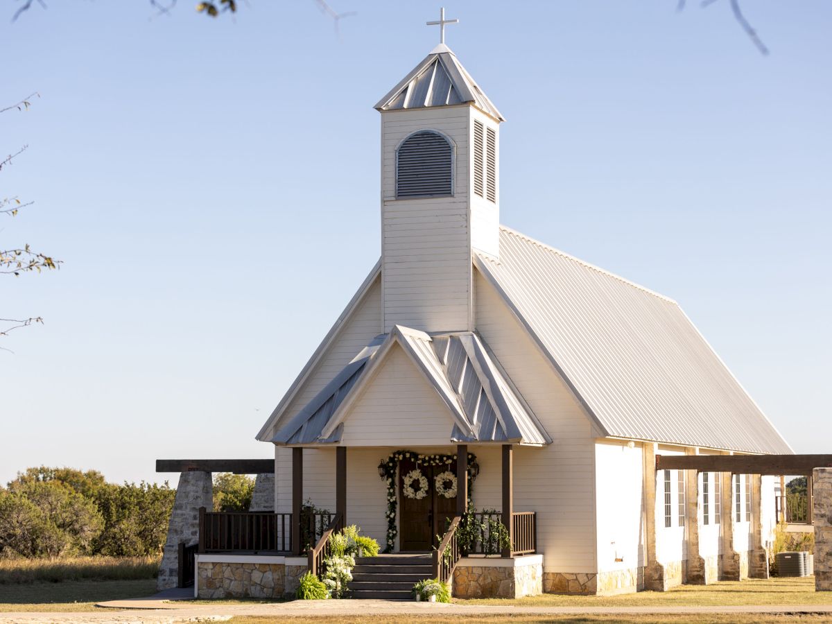 A quaint white church with a bell tower and cross, set in a rural landscape under a clear blue sky.