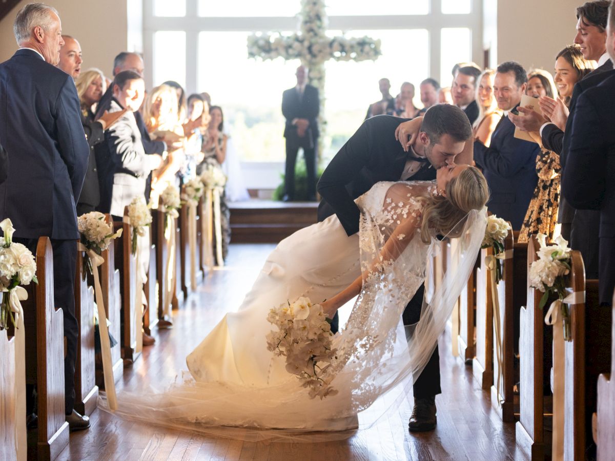 A couple shares a romantic kiss in the aisle during their wedding ceremony, surrounded by guests joyfully celebrating.