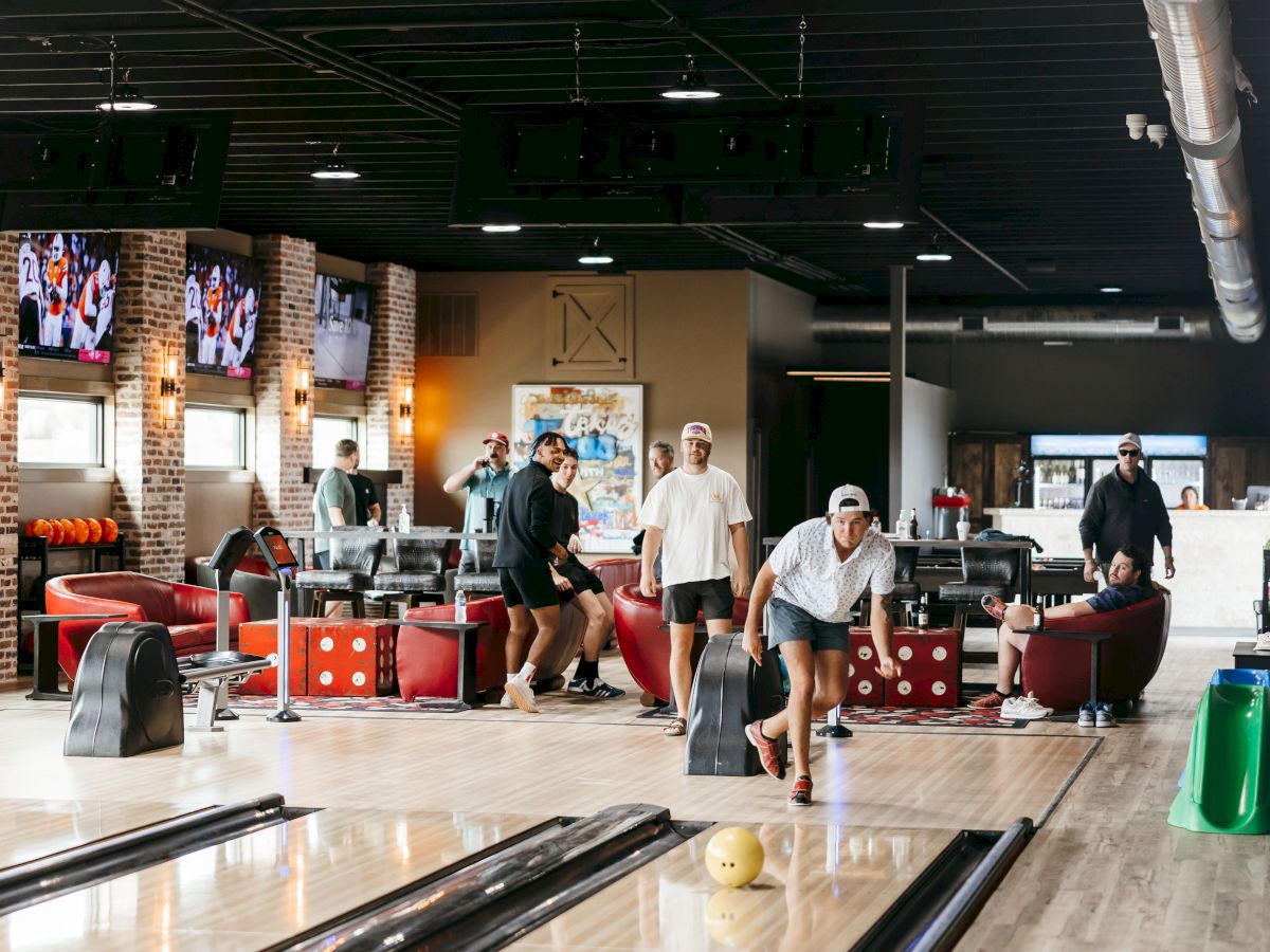A group of people is bowling in a modern alley with comfortable seating, large TVs, and brick accent walls.