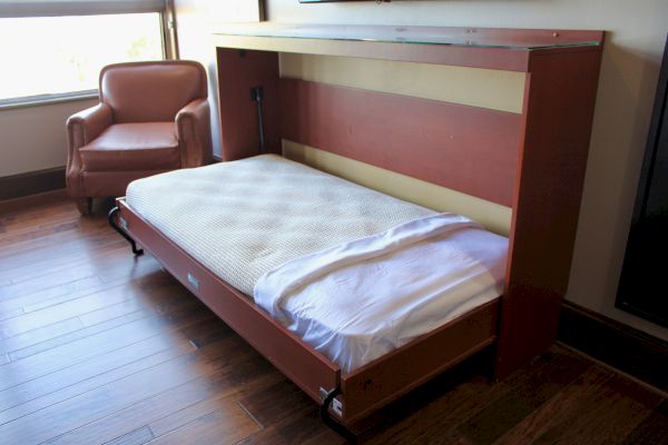 A partially unfolded Murphy bed in a room with hardwood flooring and a brown armchair near a window.