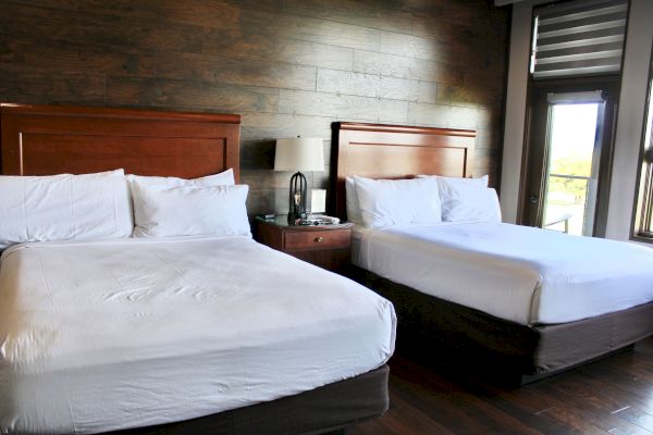 The image shows a hotel room with two neatly made beds, a nightstand, and a lamp, featuring wooden wall panels and a window.
