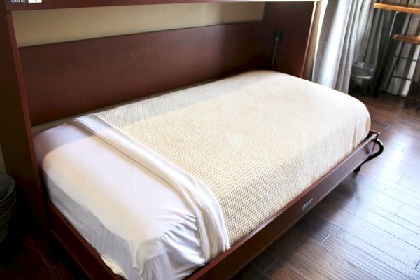 A neatly made bed with a white sheet and beige blanket in a wooden frame is shown on a hardwood floor.