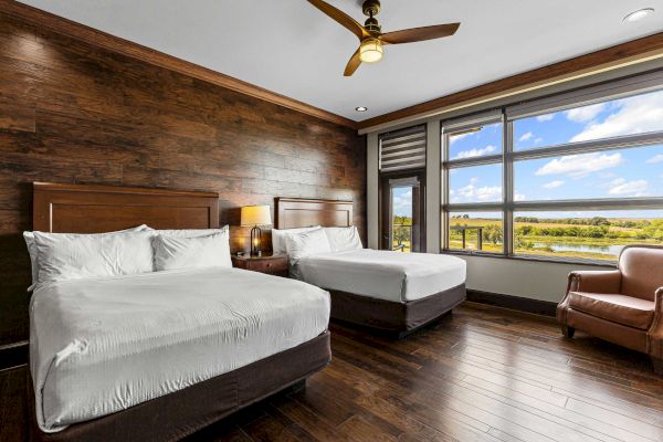 The image shows a hotel room with two beds, a wooden wall, a ceiling fan, large window with scenic view, and an armchair.