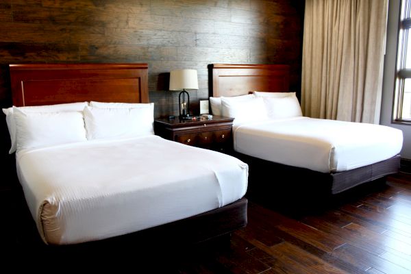 The image shows a hotel room with two large beds, a bedside table with a lamp, and curtains, all with a wooden floor and wall backdrop.