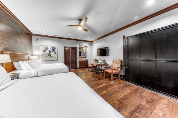 A spacious hotel room with two beds, a ceiling fan, TV, wooden floor, and modern decor featuring a painting, chairs, and a cabinet.