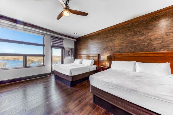 A modern hotel room with two beds, wooden flooring, a ceiling fan, large windows with a view of water, and a warm, inviting ambiance.