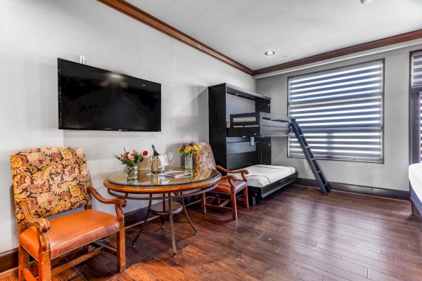 A room with a TV, table with chairs, flowers, wine, and a bunk bed next to a window with striped blinds.