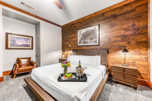 A cozy bedroom with a wooden accent wall, a bed with a breakfast tray, two paintings, a leather chair, and a nightstand with a lamp.