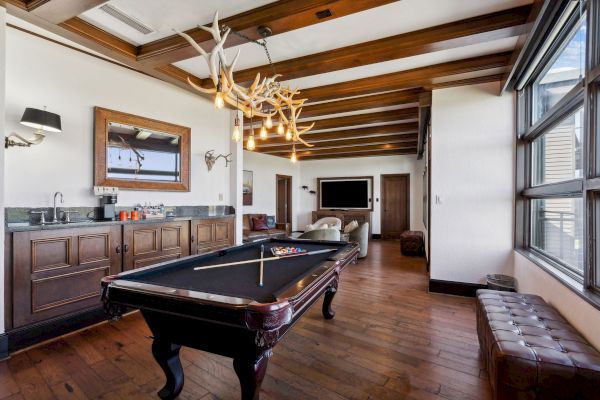 The image shows a game room with a pool table, antler chandelier, sofa, large window, and built-in bar area with wooden cabinetry.