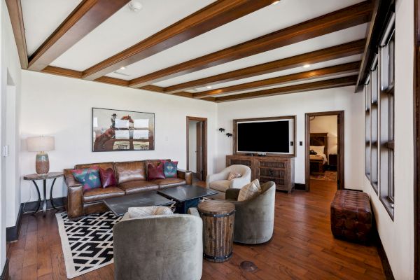The image shows a cozy living room with a leather sofa, armchairs, TV, and wooden beams on the ceiling.