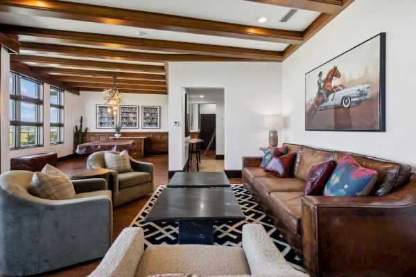 A cozy living room features leather sofas, armchairs, a stylish rug, a modern art piece, and a wood-beamed ceiling with ample natural light.